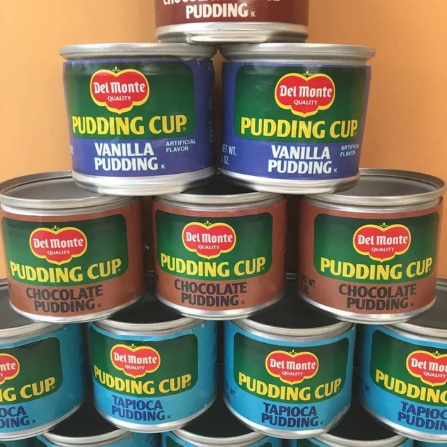 A pyramid stack of Del Monte pudding cups in three flavors: vanilla, chocolate, and tapioca, with colorful green, blue, and brown labels against a light background.
