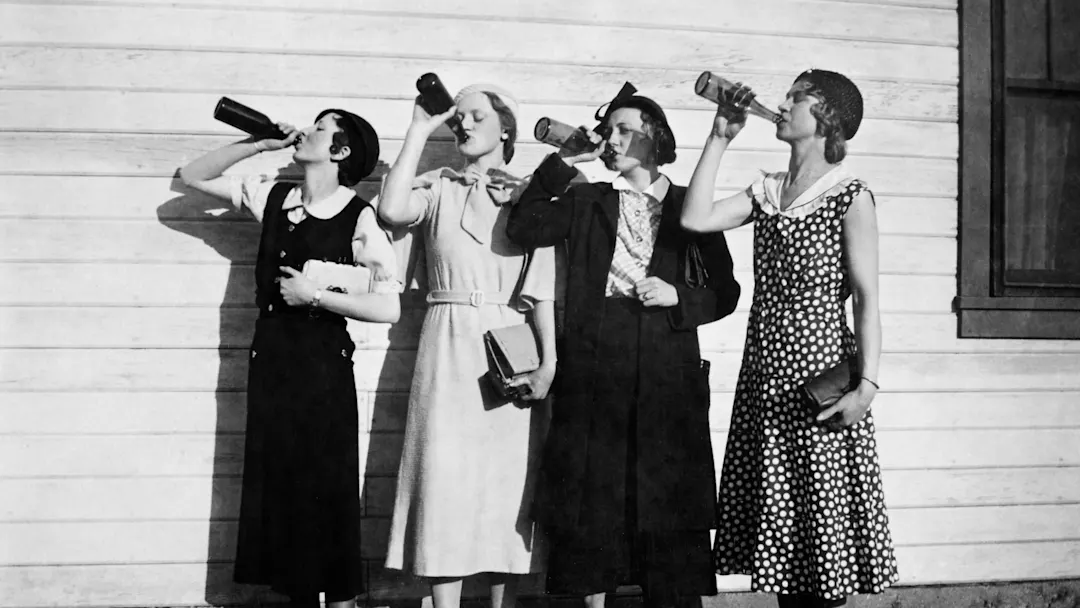 Four women in vintage dresses stand in a row outdoors against a wooden wall, each drinking from a bottle and holding books or bags, with sunlight casting shadows behind them.