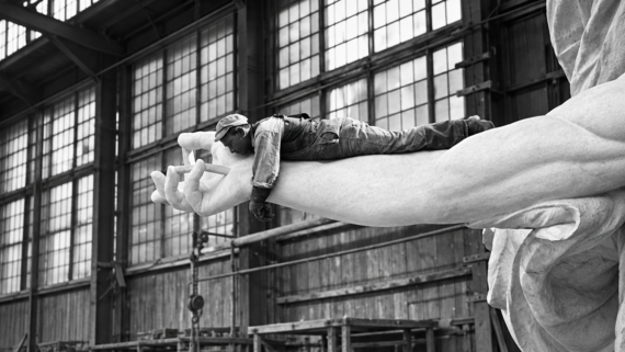 A person in work clothes lies face down on the outstretched arm of a large statue inside an industrial building full of windows and scaffolding.
