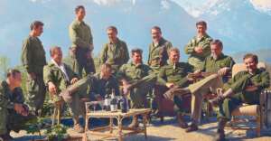 A group of thirteen men in olive military uniforms relax and pose outdoors with mountains in the background. Some are seated around a table with bottles and glasses, while others stand, all appearing to be in good spirits.
