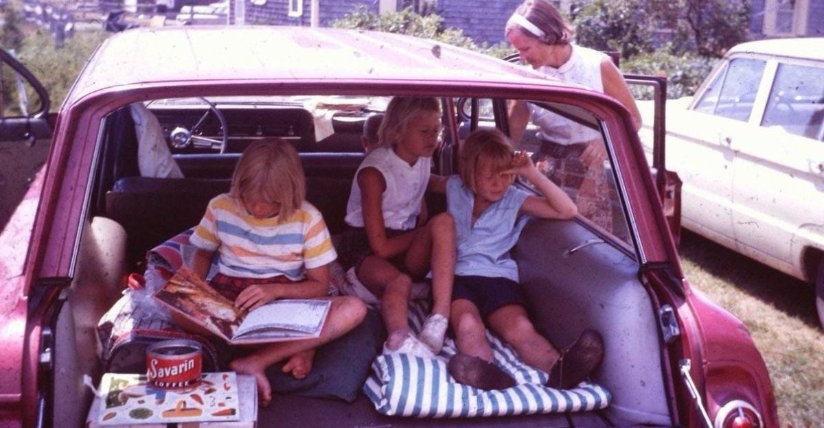 Three children sit in the open trunk of a vintage car, reading and relaxing, while an adult woman stands nearby with the car door open. They appear to be preparing for or taking a break during a trip.