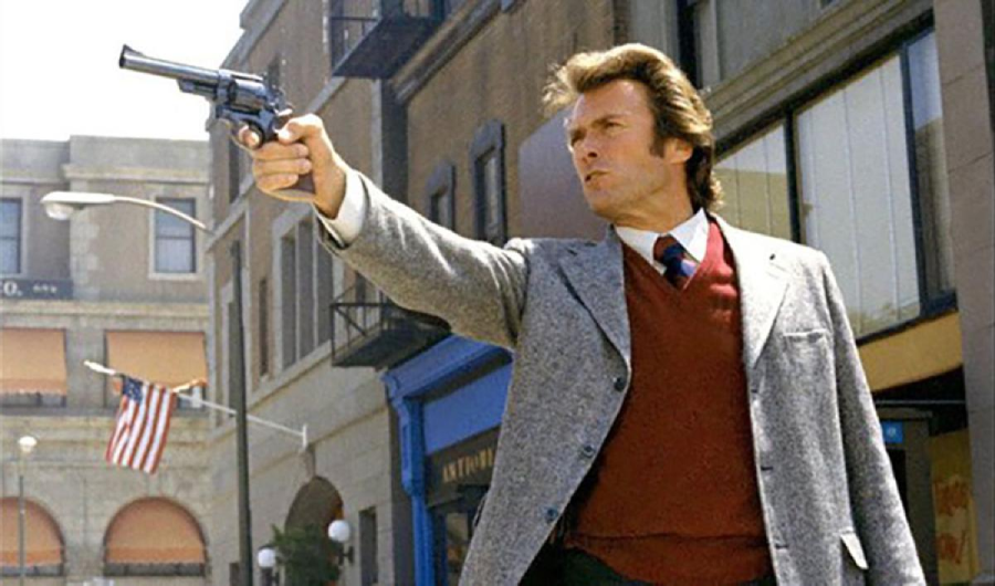 A man in a gray blazer and red sweater aims a revolver on a city street with buildings and an American flag in the background.