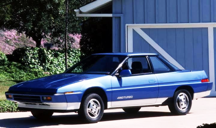 A blue two-door coupe with “4WD TURBO” written on the door is parked in a driveway in front of a blue house with white trim, surrounded by greenery on a sunny day.
