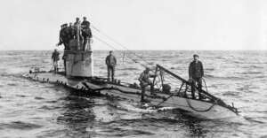 A group of sailors stands on the deck and conning tower of a surfaced World War I-era submarine in open water, with cables and equipment visible along the length of the vessel.