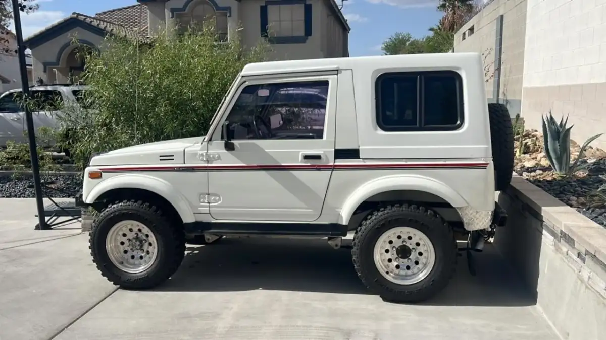 A white two-door off-road vehicle with large tires and black and red side stripes is parked on a concrete driveway beside a house and a garden with desert plants.