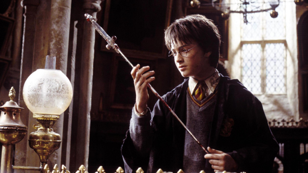 A young person wearing glasses and a school uniform robe examines a long, ornate sword in an old, dimly lit room with tall windows and gothic decor.