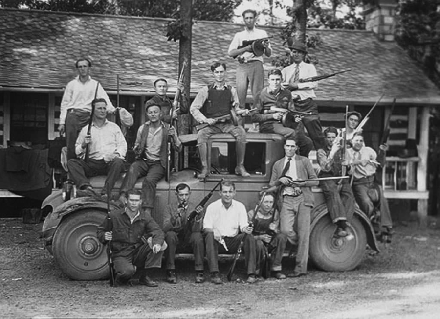 A group of men, many holding rifles and shotguns, pose around and on top of an old car in front of a rustic building. Most are wearing hats and work clothes, suggesting an early 20th-century rural setting.