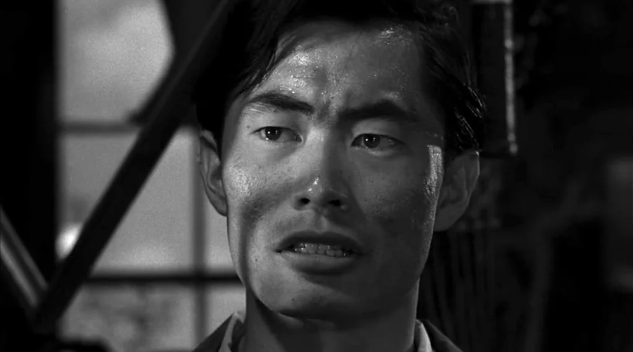 Black-and-white close-up of a man with an anxious expression, furrowed brows, and slightly parted lips, standing in front of a window with light streaming in.