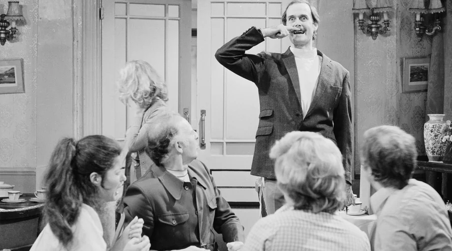 A man in a suit stands in a room making a funny face and gesture, while four seated people watch him and another person walks away in the background. The setting appears to be a restaurant or cafe.