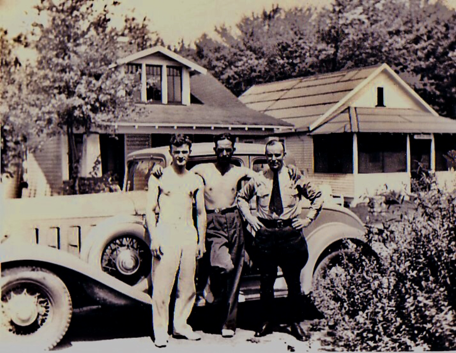 Three men stand in front of a vintage car parked outside two houses, with trees in the background. The photo has a sepia tone, suggesting it is from the early to mid-20th century.