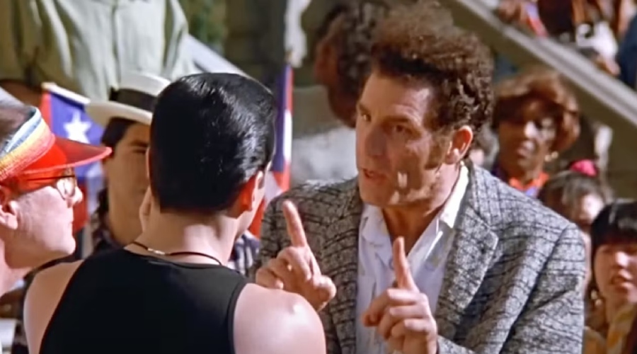 A man in a patterned jacket gestures with both index fingers raised while talking to another man in a black tank top, surrounded by a crowd of people outdoors.