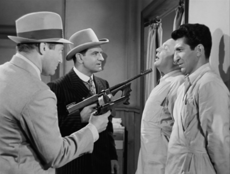 A man in a fedora points a tommy gun at two men pressed against a wall, while another man in a striped suit and hat looks on; the scene appears tense and dramatic, set in a vintage black-and-white film.