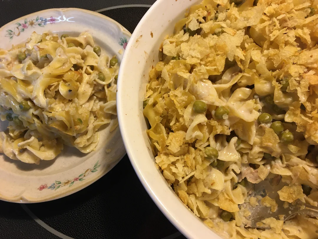 A casserole with egg noodles, peas, and a creamy sauce topped with crushed potato chips is served on a floral plate, next to the main baking dish.