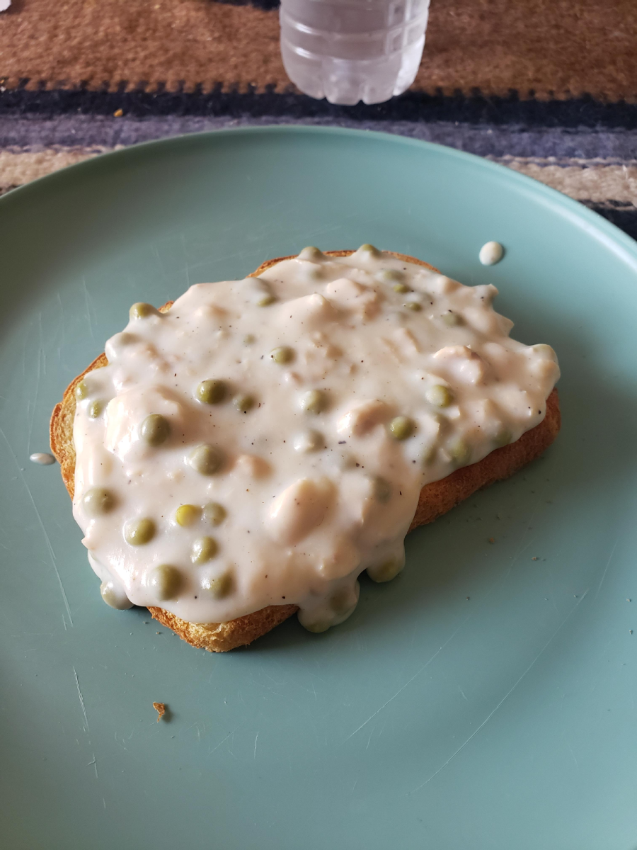 A slice of toast topped with a creamy mixture containing peas and possibly small pieces of meat, served on a light blue plate. A partially visible water bottle is in the background.