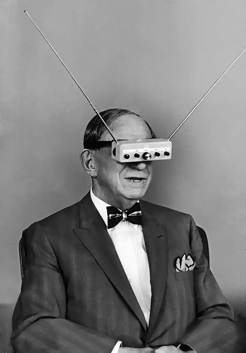 A man in a suit and bow tie sits, wearing a boxy device with buttons and two long antennae over his eyes, giving the appearance of futuristic or experimental eyewear.