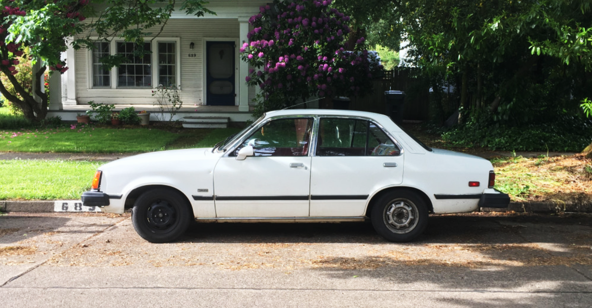 A white, older model sedan is parked on a residential street in front of a house with a porch and blooming purple flowers. Trees and greenery surround the area, giving a peaceful neighborhood feel.