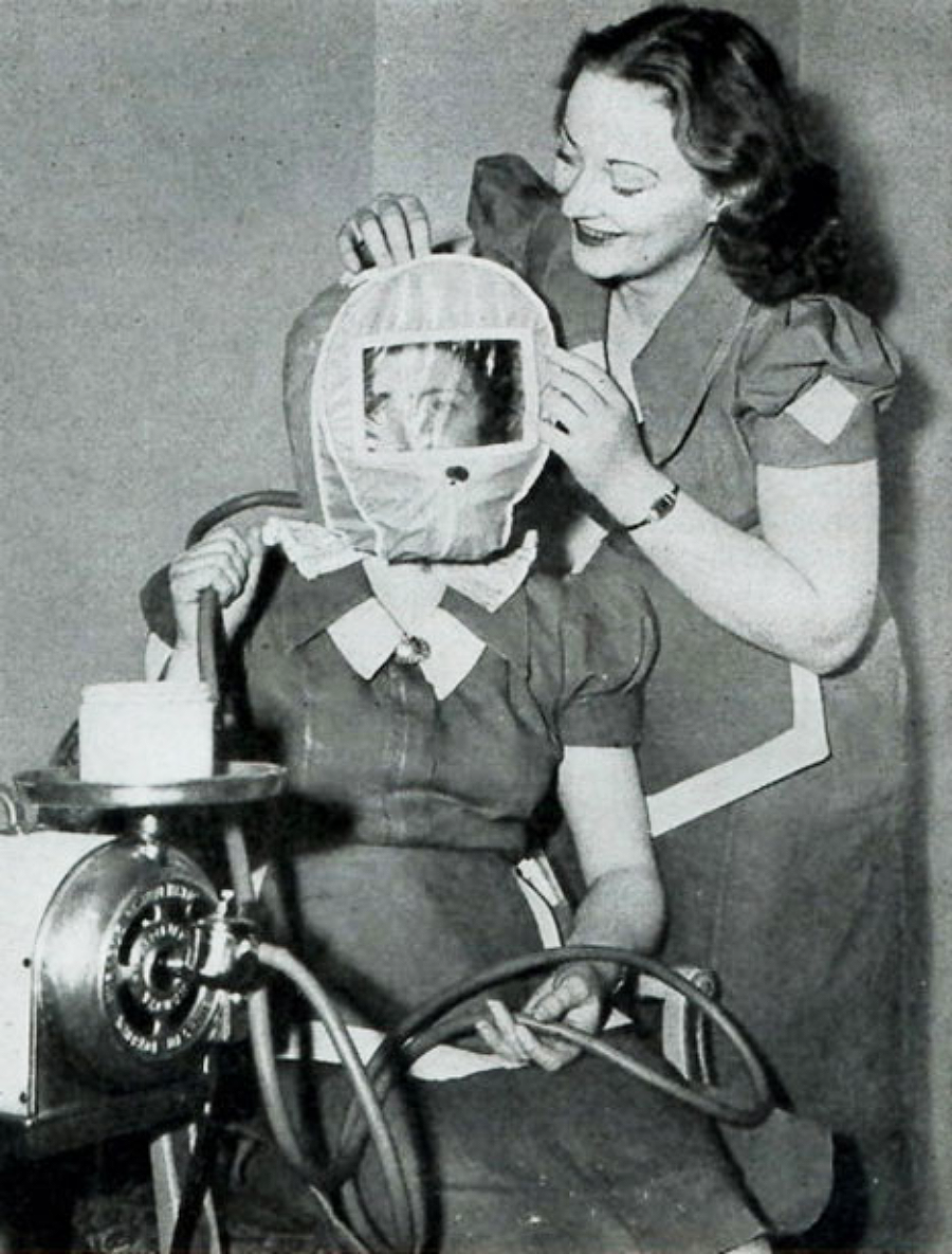 A woman places a transparent protective hood over another woman's head, who is seated and holding hoses attached to a nearby machine. Both women are wearing matching dresses with white collars. The setting appears to be indoors.