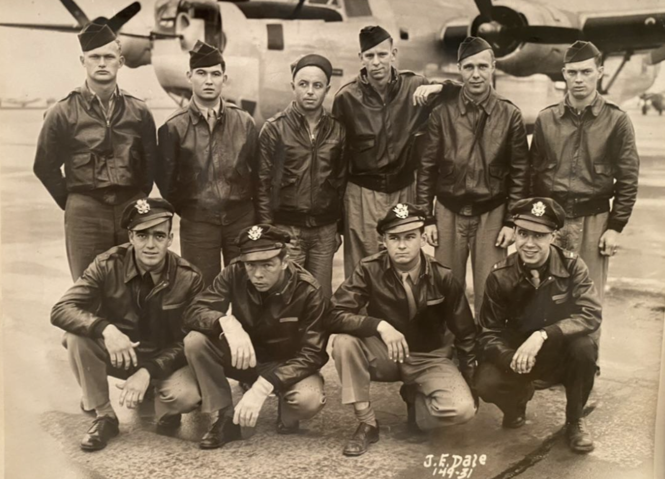 A group of ten men in military uniforms and jackets pose in front of a large propeller airplane on an airfield. Four are kneeling in front, and six are standing behind them. The photo is in black and white.