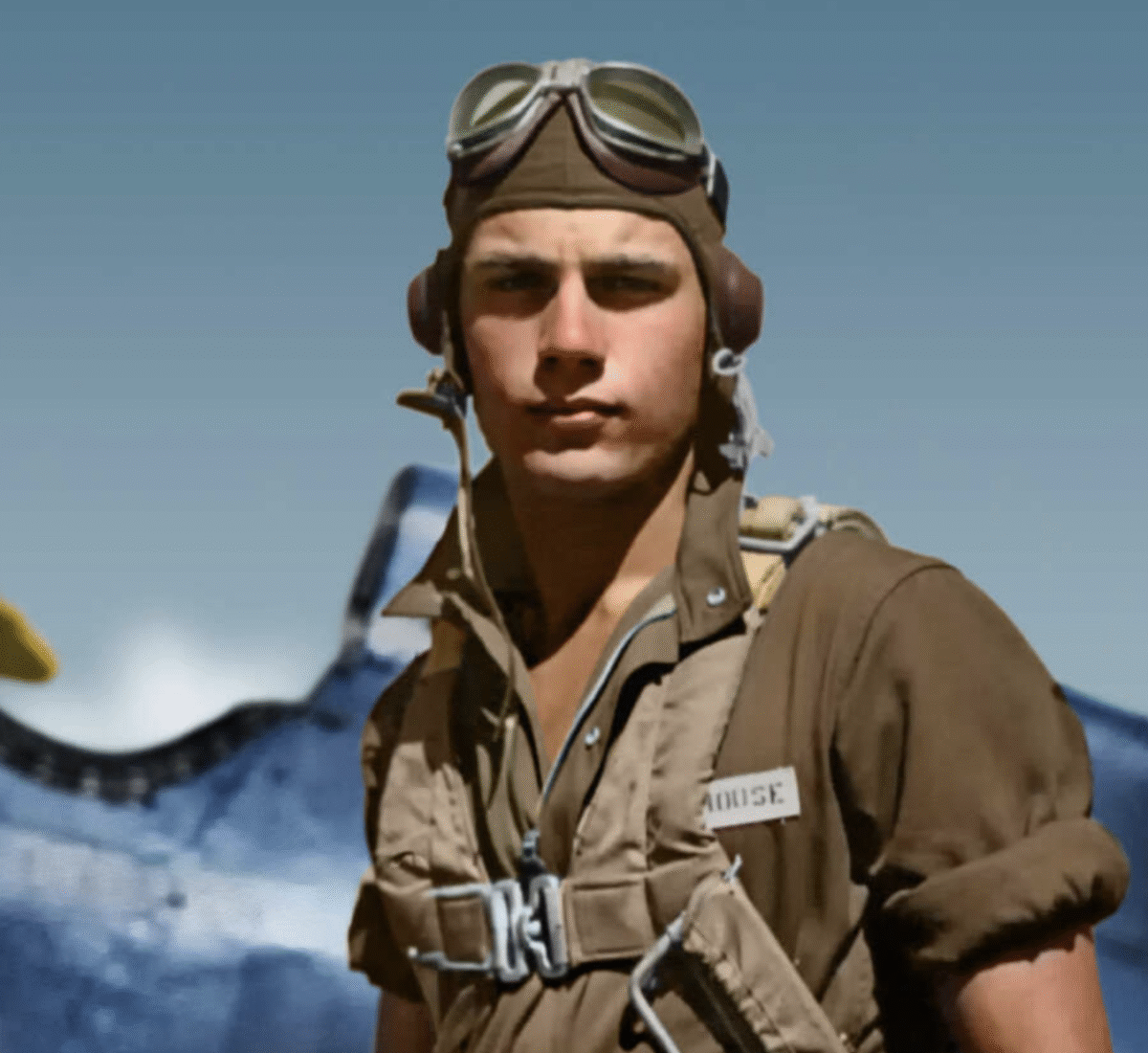 A young World War II pilot wearing goggles, headphones, and a brown flight suit stands in front of a vintage aircraft, looking at the camera with a serious expression.