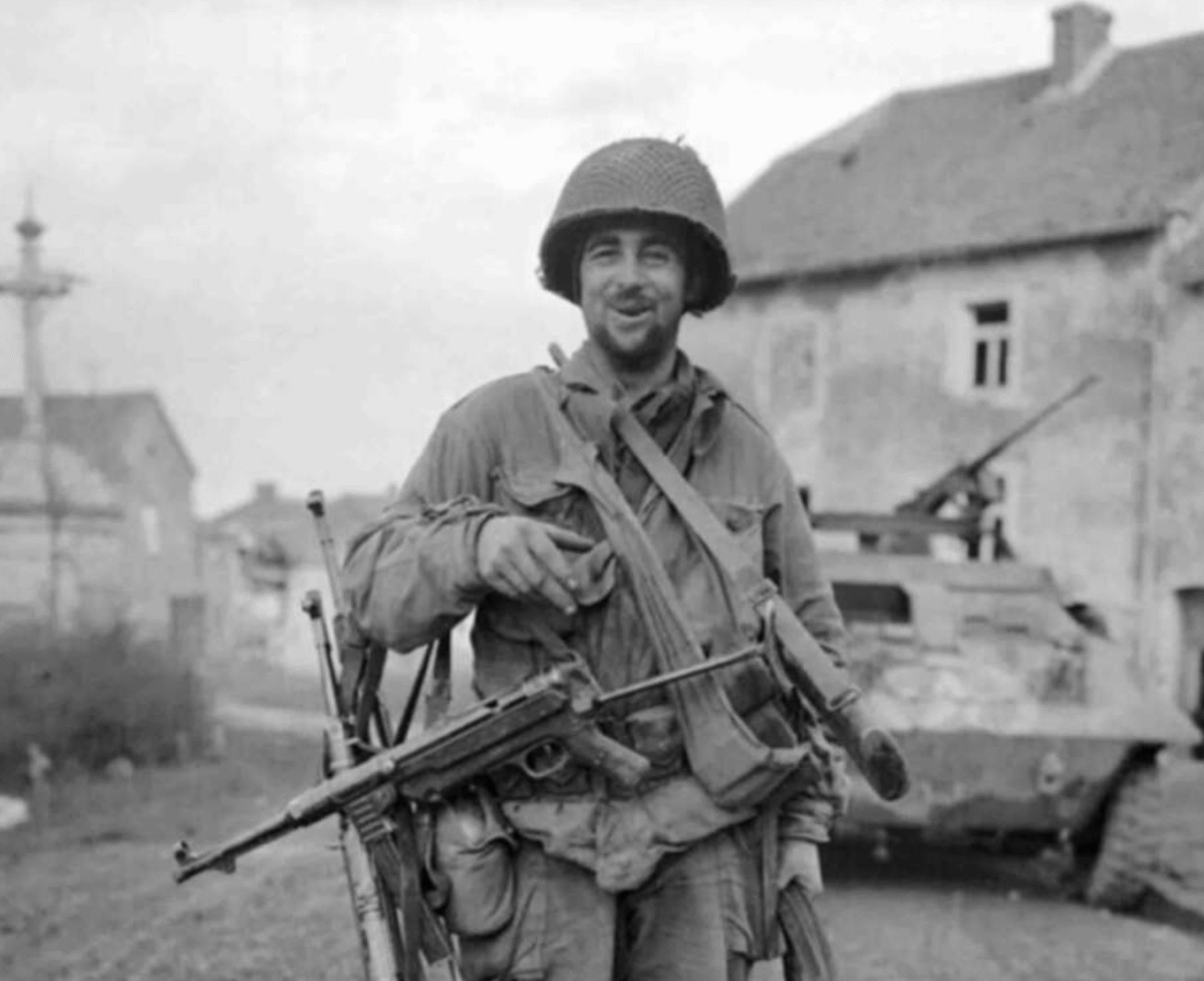 A soldier in World War II uniform smiles at the camera, holding a weapon and standing on a street with a damaged building and a military vehicle in the background.