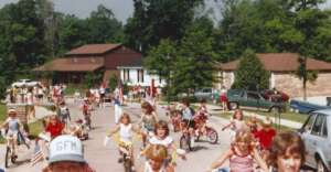 Children ride decorated bicycles in a neighborhood parade, many wearing red, white, and blue outfits. Trees and houses with parked cars line the street on a sunny day.