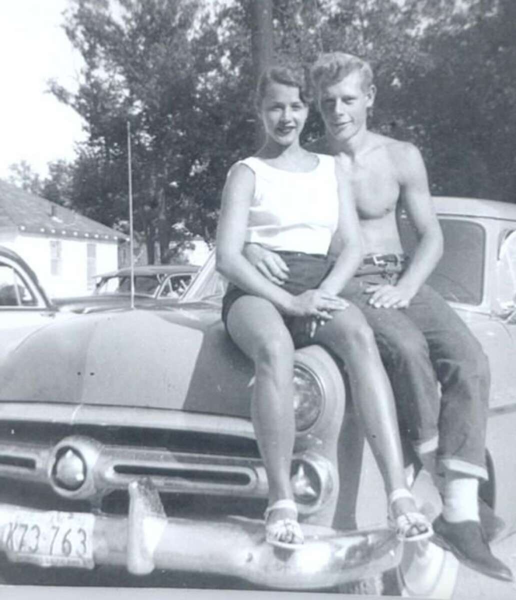 A young woman and a shirtless young man sit together on the hood of a vintage car, smiling at the camera. Trees and other parked cars are visible in the background. The image appears to be from the 1950s or 1960s.