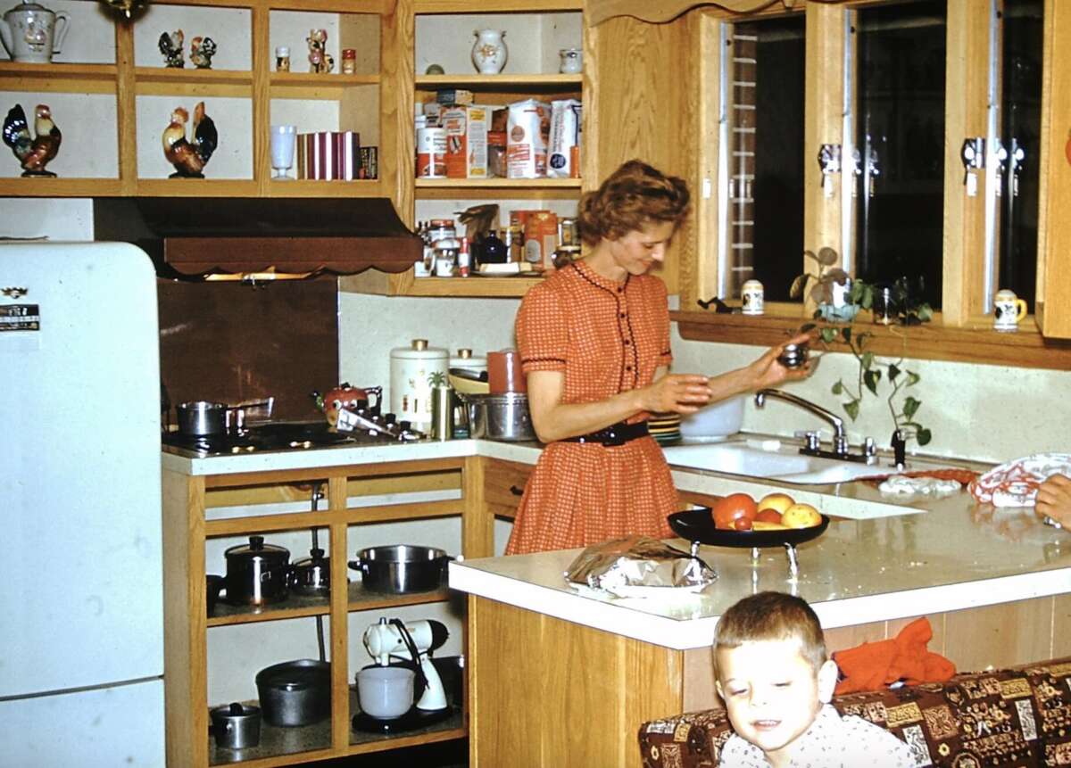 A woman in an orange dress prepares food at a kitchen counter, while a child sits nearby. The kitchen has wooden cabinets, a white refrigerator, and shelves with dishes and canned goods. A bowl of fruit is on the counter.