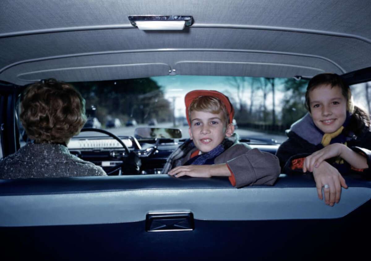 Two children sit in the back seat of a car, smiling at the camera, while an adult drives. The scene is viewed from inside the car, showing the road and trees through the front windshield.