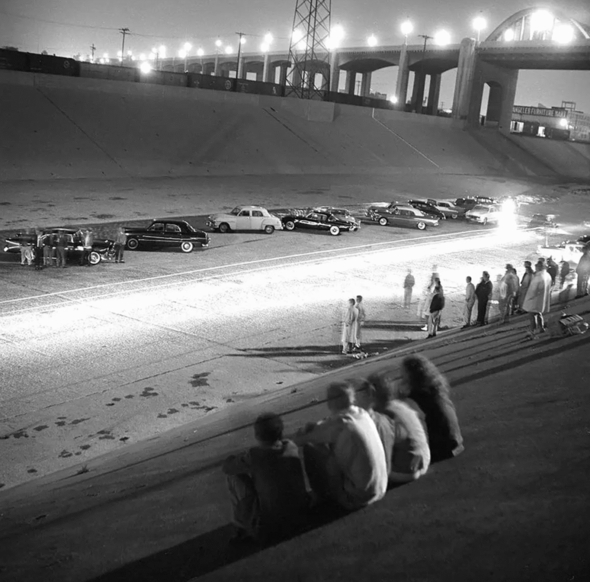 A black-and-white photo shows people watching a nighttime car race on a wide, empty road under an overpass. Classic cars are parked along the roadside, and spectators sit on concrete steps in the foreground.