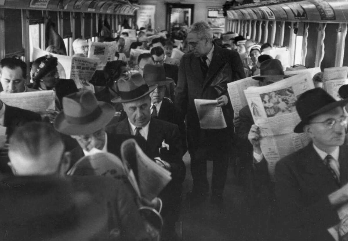 Passengers on a vintage train, mostly men in suits and hats, sit and stand while reading newspapers. The scene is crowded and everyone appears absorbed in their papers.