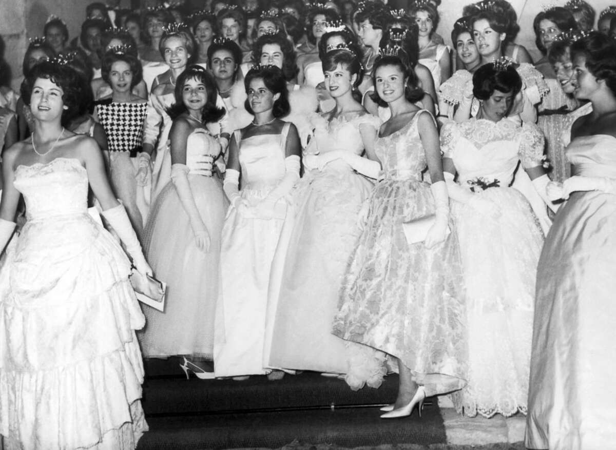 A large group of young women in formal gowns and gloves pose together, many wearing tiaras. They are smiling and standing on a staircase, appearing to be at a formal event or ball from a past era.