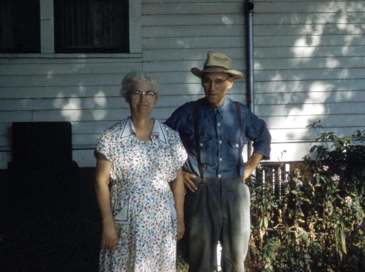 An elderly woman in a floral dress and glasses stands next to an elderly man in suspenders, a hat, and glasses. They are outdoors in front of a white house with flowers and shrubs nearby.