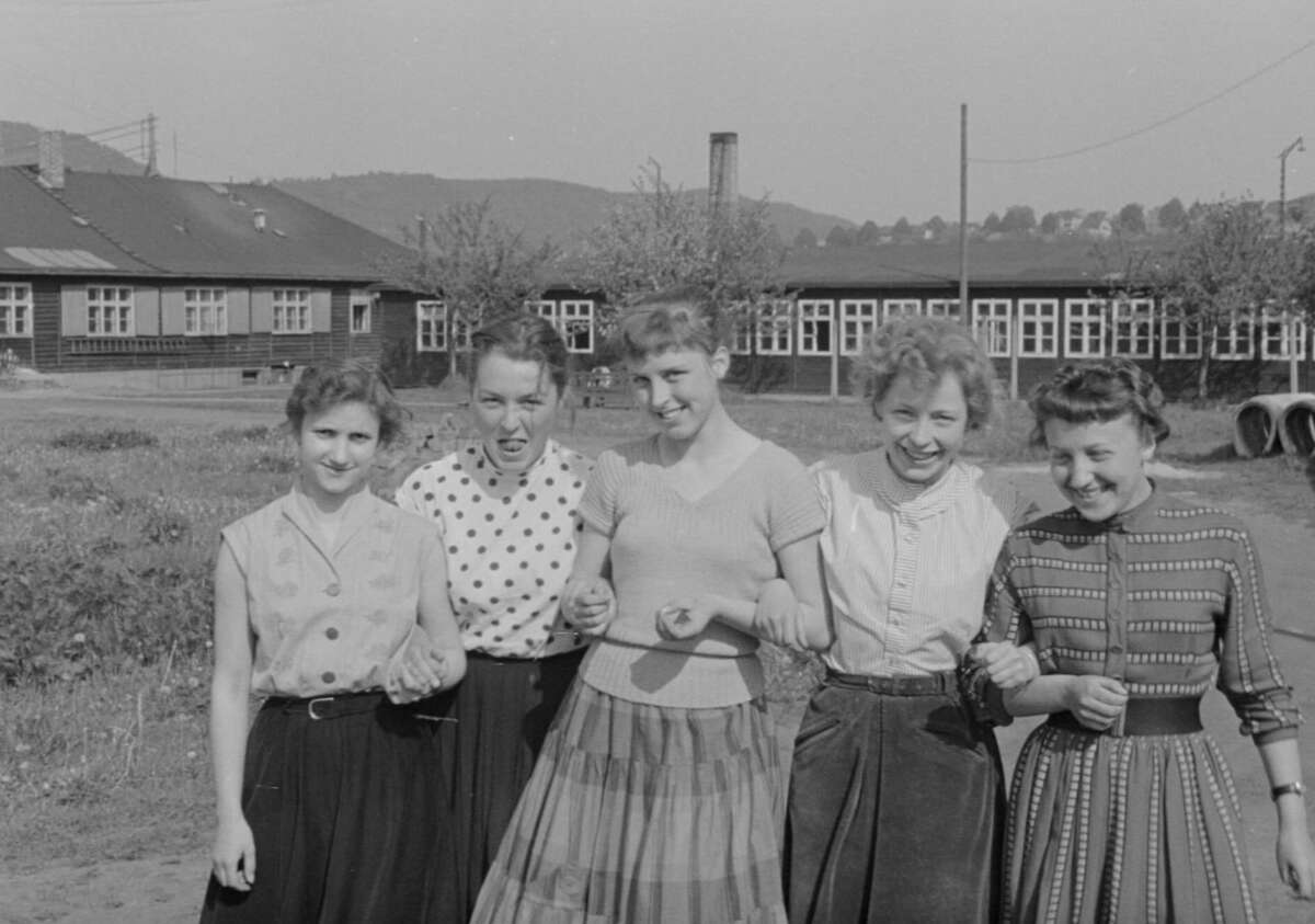Five young women stand outdoors in a row, linking arms and smiling. They wear mid-20th century skirts and blouses. Behind them are buildings, grassy fields, and trees, with hills visible in the distance.