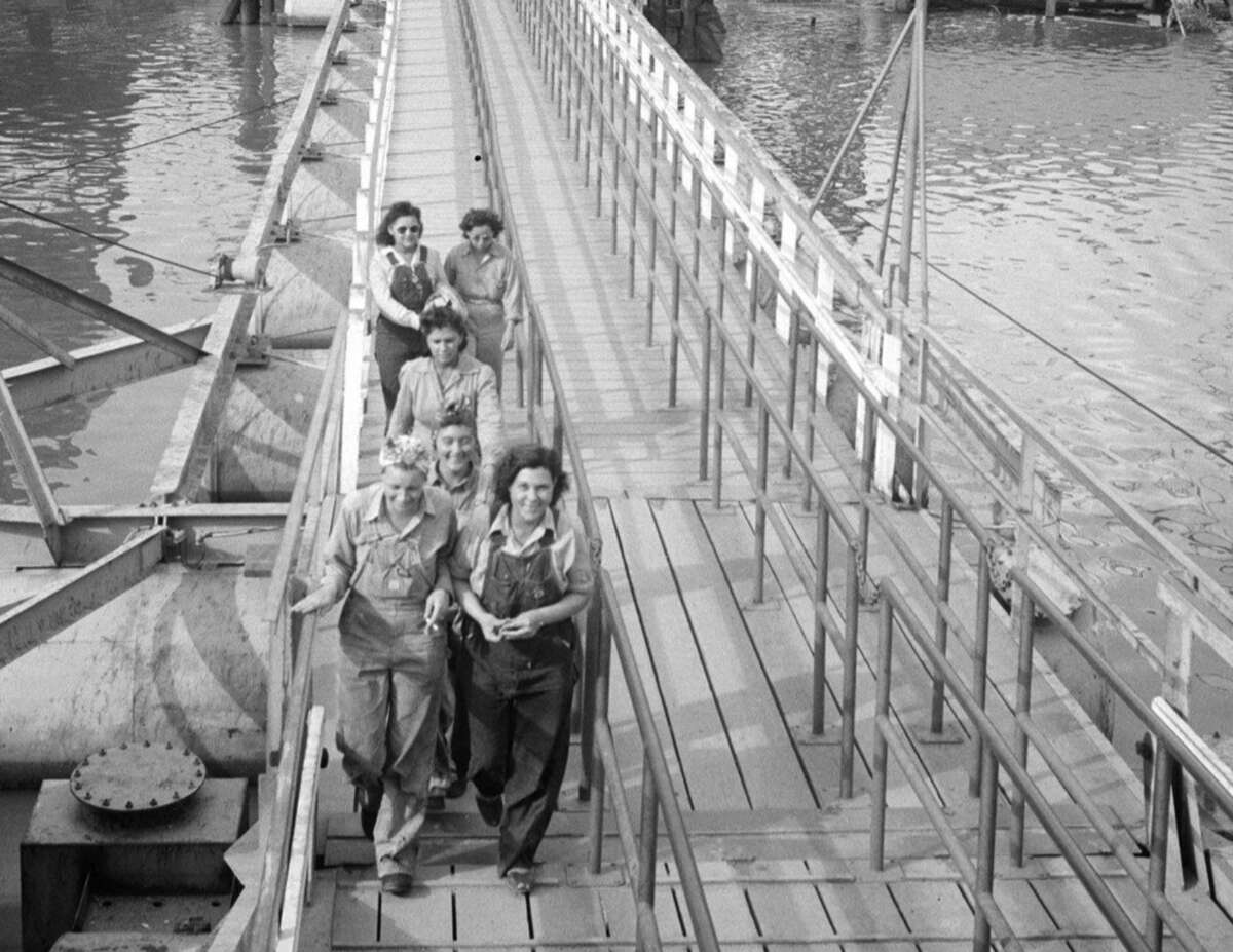 A group of women, some in work overalls, walk together across a floating pontoon bridge over a body of water, smiling and chatting as they head toward the camera.
