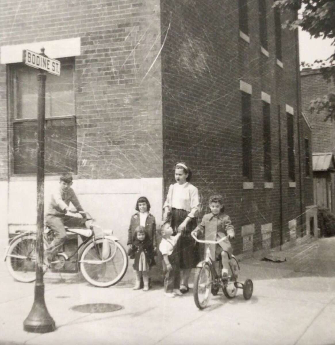 A vintage black-and-white photo shows a woman and three children on a city sidewalk near a street sign reading "Bodine St." A man sits on a motorbike, and a child rides a tricycle. Brick buildings line the street.