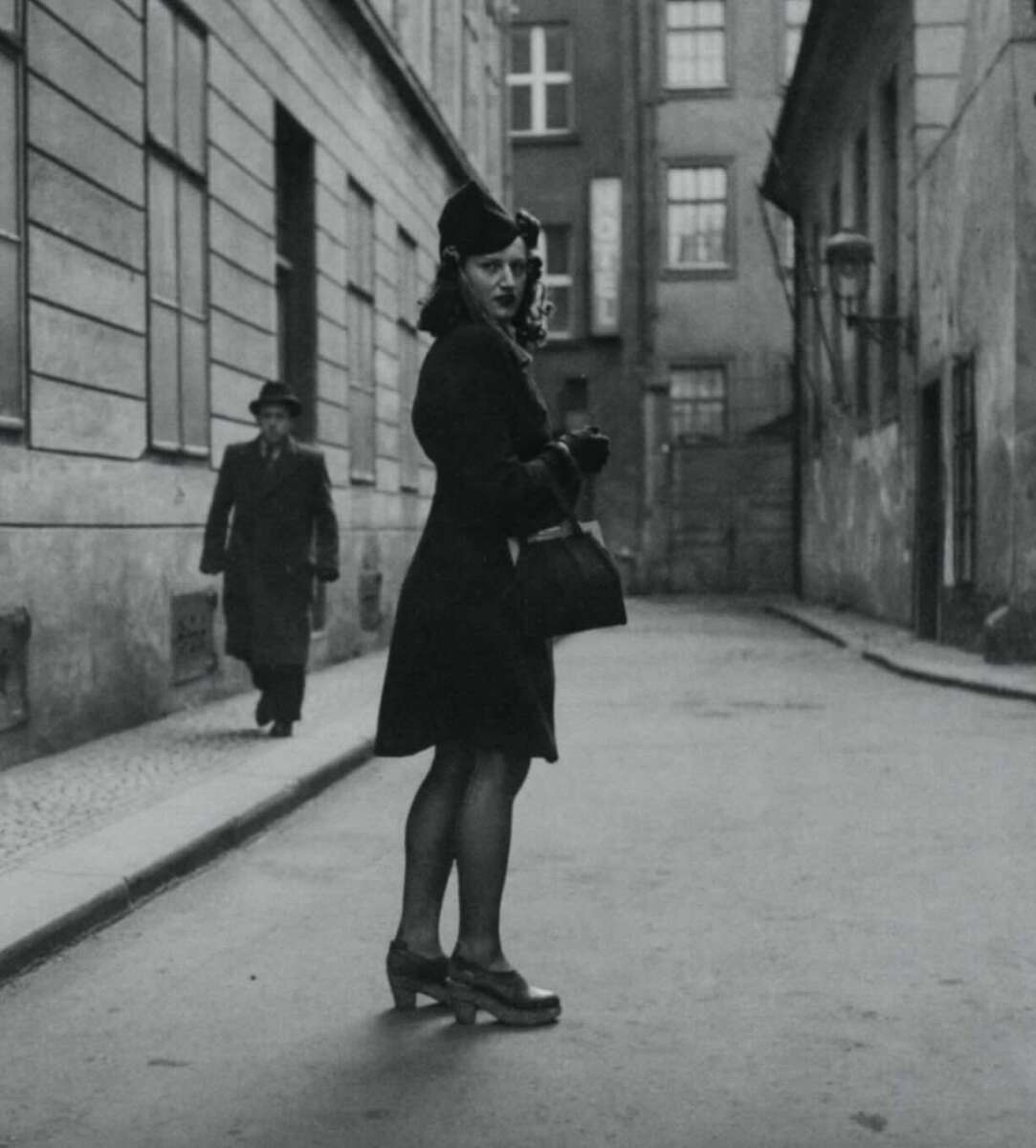 A woman in a dark coat, hat, and heels stands on a deserted city street, looking over her shoulder. A man in a long coat and hat walks in the background. The scene appears to be from an earlier era.