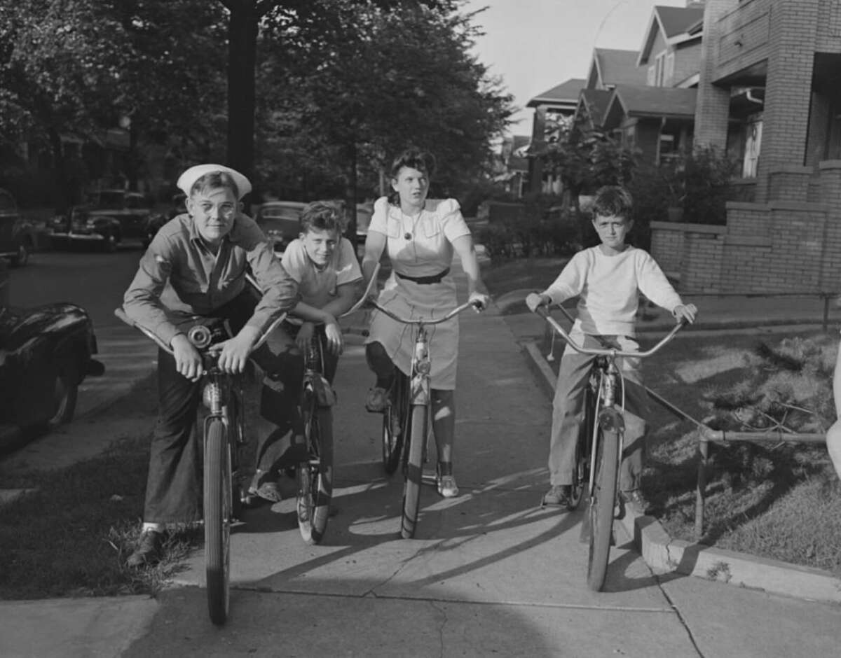 Four children on bicycles pose on a residential sidewalk in a suburban neighborhood, with trees, parked cars, and houses in the background. The photo appears to be from the mid-20th century.