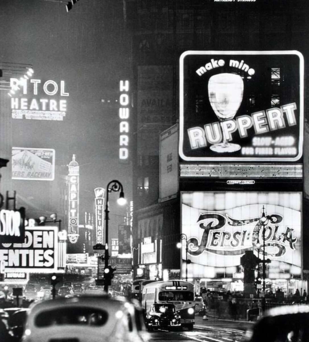A busy city street at night in the mid-20th century, with cars, buses, and people, illuminated by large neon signs and billboards advertising brands like Ruppert and Pepsi-Cola.