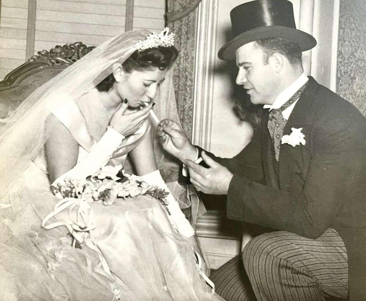 A bride in a wedding dress and veil is lighting a cigarette with the help of a man in a top hat and suit, who holds a lighter for her. They appear to be seated indoors, and the scene is in black and white.
