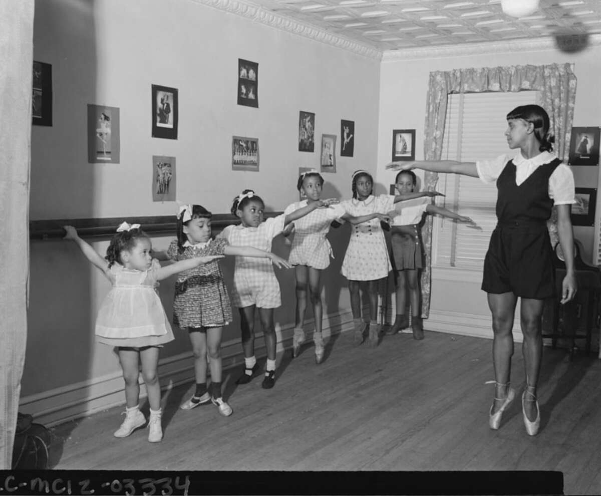 A ballet teacher in pointe shoes leads a group of young children who are lined up at a barre in a dance studio, each with one arm extended, practicing a ballet position. The room has framed photos on the walls and a window with curtains.