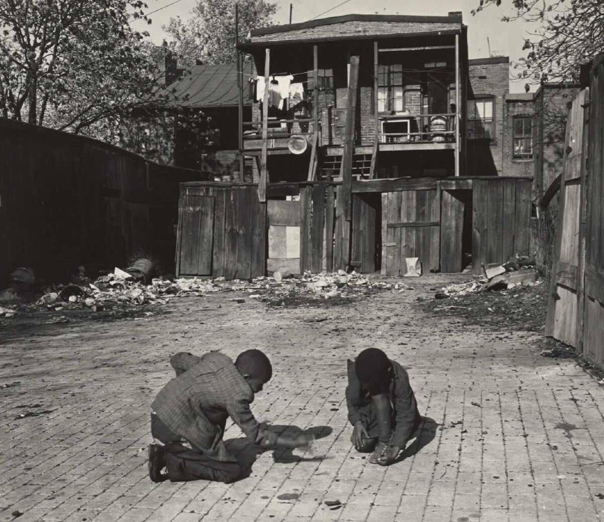 Two children crouch on a brick-paved alley, playing a game. Behind them are wooden fences, scattered debris, and a two-story building with balconies, stairs, and laundry hanging outside.