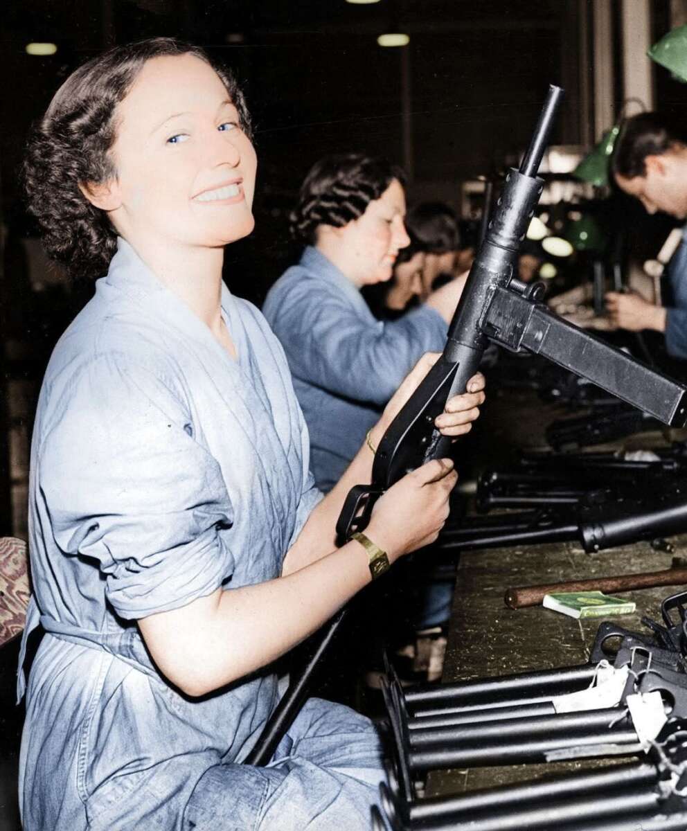 A woman in blue overalls smiles while assembling a submachine gun at a factory workbench, with other workers and gun parts visible in the background.