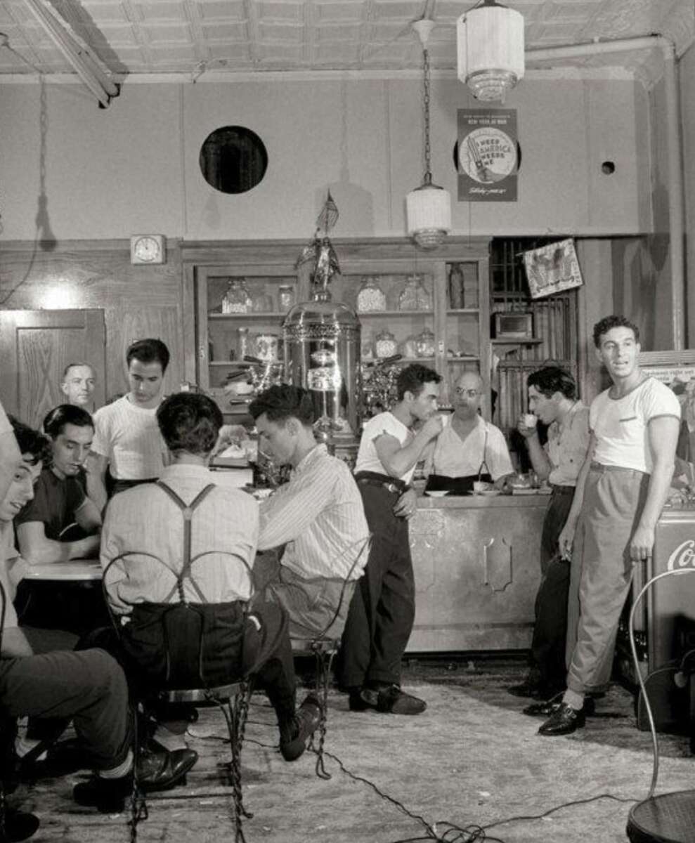 A group of men in casual 1950s attire gather in a diner or soda shop, some sitting at tables and others standing near the counter, under vintage lighting and decor.