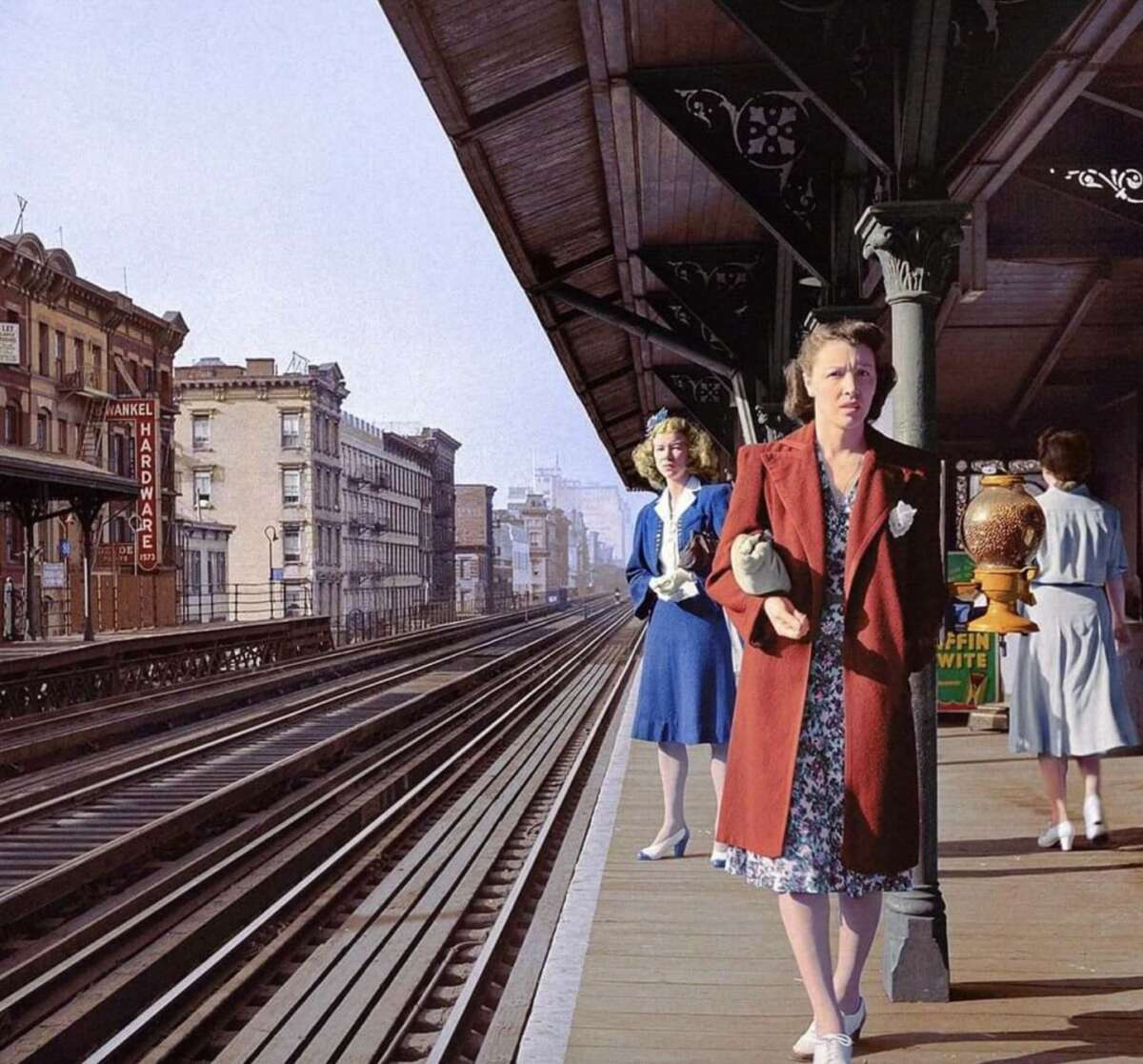 Three women stand on an elevated train platform in a city. One woman in a red coat holds a purse and looks ahead, while another in blue stands near the edge, and the third, in gray, faces away. Old buildings line the street below.