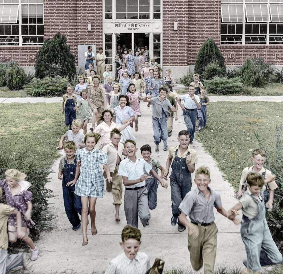 A large group of joyful children run out of the entrance of a brick school building marked "BUCKLIN PUBLIC SCHOOL," smiling and laughing on a bright day.