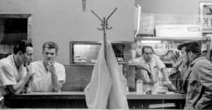 Black-and-white photo of two men talking behind a diner counter as another man in a hat sits smoking; a staff member stands behind the counter. A coat rack and hanging coat are in the foreground, dividing the scene.