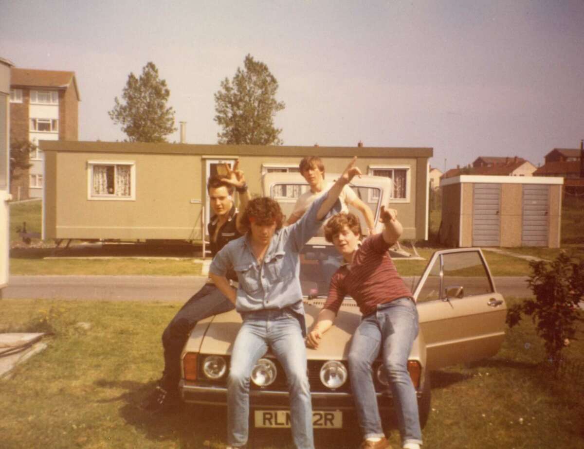 Four young men pose playfully on the hood of a tan car with the door open, outside a green mobile home on a sunny day, with houses and trees in the background.