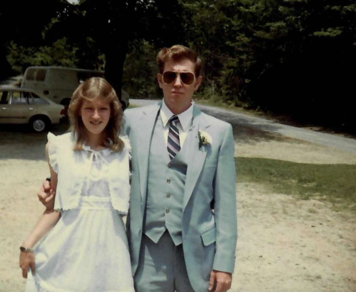 A young woman in a white dress stands next to a man wearing a light blue suit, tie, and sunglasses. They are outdoors on a sunny day, with trees and parked cars in the background.