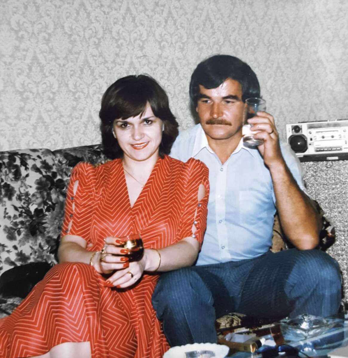 A woman in a red dress and a man in a light blue shirt sit close together on a floral-patterned sofa, both holding drinks, with vintage wallpaper and a radio in the background.