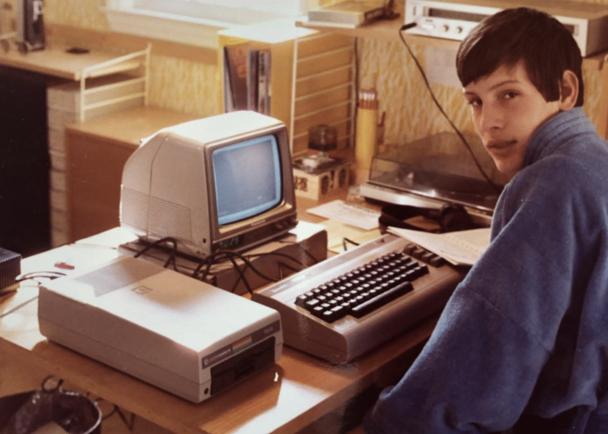A young person in a blue sweater sits at a desk with a vintage computer setup, including a small CRT monitor, keyboard, and floppy disk drive, in a room with warm lighting.